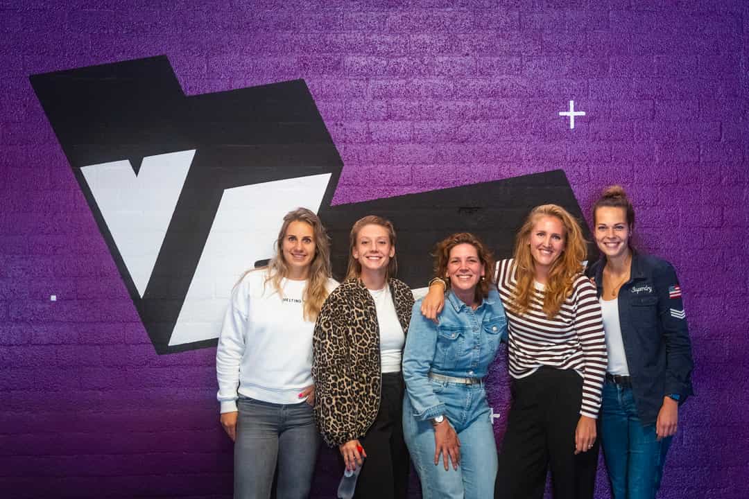 A group of women posing against a purple wall with a big VRcafe logo on it after their bachelor's party outing